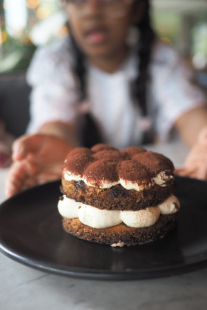 A child holds out a dessert with layers of cream and cake in a cafe setting