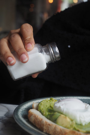 Person adding salt to avocado toast at cafeの写真素材
