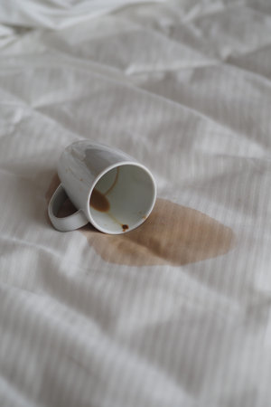 Coffee cup spills on a white bed sheet in morning light