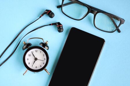 Black glasses, cellphone, alarm clock and headphones on blue background composition. Flat lay and top view photoの写真素材