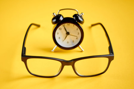 Black glasses and alarm clock on yellow background composition, eyeglasses. Flat lay and top view...