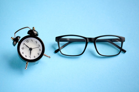 Black glasses and alarm clock on blue background composition. Flat lay and top view photo