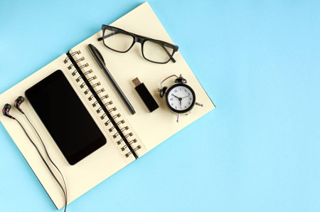 Black glasses, cellphone, paper notepad, alarm clock and headphones on blue background composition....