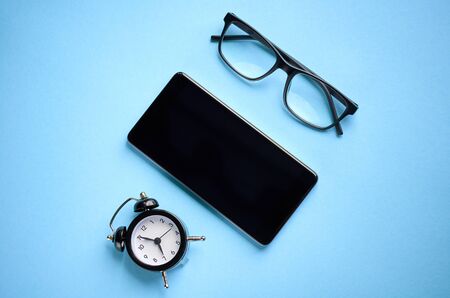 Black glasses, alarm clock and cellphone on blue background composition. Flat lay and top view photoの写真素材