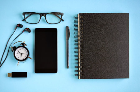 Black glasses, cellphone, paper notepad, alarm clock and headphones on blue background composition....