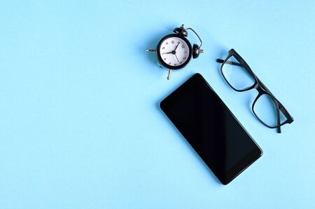 Black glasses, alarm clock and cellphone on blue background composition. Flat lay and top view photoの写真素材
