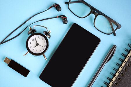 Black glasses, cellphone, paper notepad, alarm clock and headphones on blue background composition. Flat lay and top view photoの写真素材