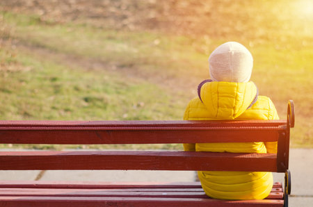 Child sits alone on wooden bench. Boy in yellow jacket and grey hat.の写真素材