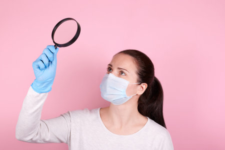 Portrait of a brunette girl in a medical mask and blue rubber gloves using magnifying lens. Isolated...