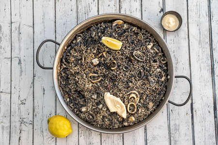 Spanish paella of black rice with squid, fish and clams with aioli sauce and lemon on white tableの写真素材