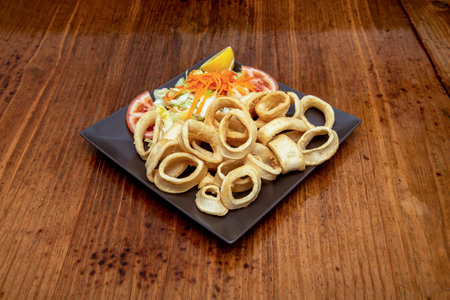 Square black plate with Spanish lid of Roman batter squid rings on dark wooden tableの写真素材