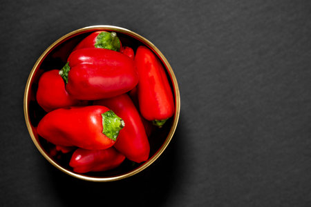 Top view of small red bell peppers inside a golden bowl on black background
