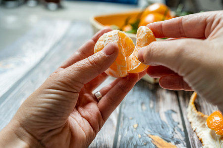 two man's hands separating a segment of ripe tangerine and in the background a basket with more mandarinsの写真素材
