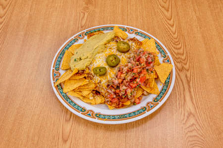 Plate of Mexican corn nachos with lots of cheese, lots of guacamole and lots of pico de gallo on white plate with border and wooden tableの写真素材