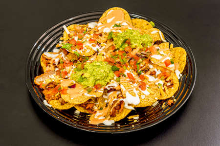 Plate of round nachos with shredded meat, guacamole, cheese in quantity with tomato mince, onion, peppers and parsley on black plateの写真素材