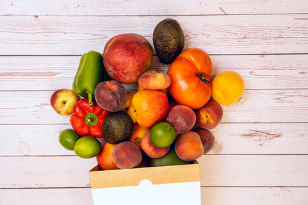Fruits and vegetables from the market pouring out of a cardboard bag on a white wooden table.の写真素材