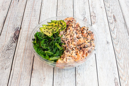Clear plastic home delivery bowl with poke bowl of salmon marinated with poppy seeds, Asian wakame seaweed salad and sliced avocadoの写真素材