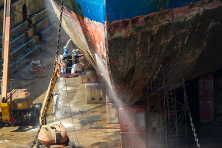 Workmen cleaning corrosion from the hull of a merchant ship with pressure sandblasting in a dry dock...