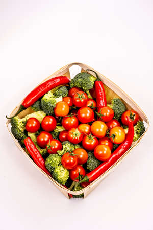 Top view image of wooden basket with bulk cherry tomatoes, broccoli pieces, red hot chili peppers on white backgroundの写真素材