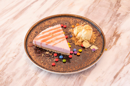 Portion of strawberry petisuis cake with assorted chocolate dragees, cookie sand and a scoop of vanilla ice cream on a brown plate and marble tableの写真素材