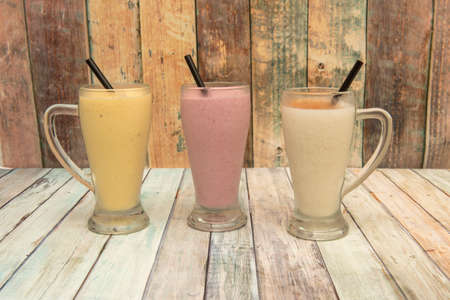 Vanilla, strawberry and mango smoothies served in glass jars with black straws on wooden tableの写真素材