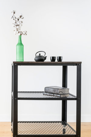 Industrial style trolley with green bottle with decorative cotton branch and black tea set with old books and white wall in the backgroundの写真素材