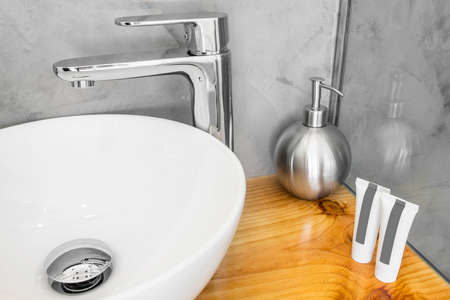 White porcelain sink with stainless steel tap on wooden countertop with soap and shampoo bottlesの写真素材