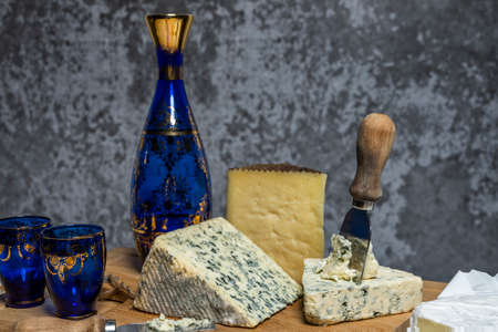 Still life with fresh and cured cheeses, cheese knives and a fine decorated blue glass bottle on a bamboo wooden tableの写真素材