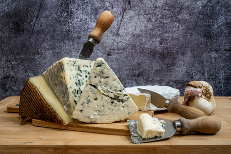 Blue cow cheese on top of cured Manchego cheese, cheese knives and head of garlic on a bamboo board