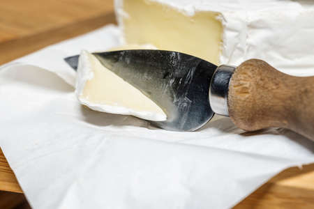 Cheese knife with a piece of brie cheese on white food paper on bamboo wood tableの写真素材