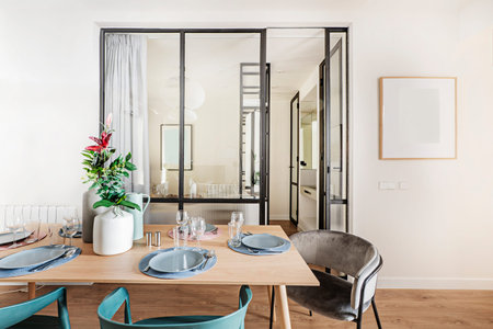 Wooden dining table with tableware set in front of black metal and glass wall of bedroom with...