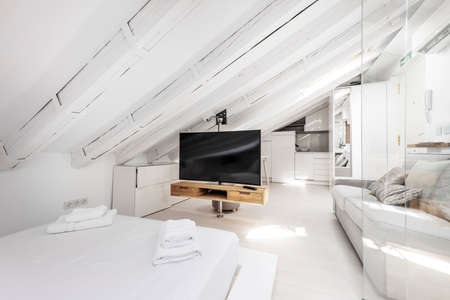 Attic apartment with exposed beams, materials and furniture painted white, flat screen TV and two-seater sofa upholstered in gray fabricのeditorial素材