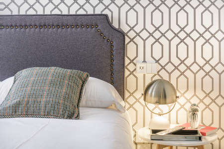 Headboard upholstered in gray fabric with vintage ocher studs, striped fabric cushion, wallpaper on the wall and lamp with steel lampshade on a white marble bedside tableのeditorial素材