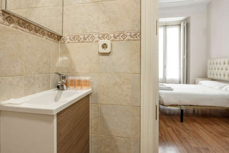Bathroom with porcelain sink on wooden furniture next to a bedroom with a double bed and a headboard upholstered in capitone fabricのeditorial素材