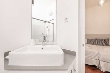 Bathroom with square white porcelain sink below mirrorless rectangular mirror next to bedroom with reddish oak flooringのeditorial素材