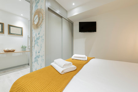 Bedroom with a double bed, towels on the bed, a mustard-colored blanket, a built-in wardrobe with gray wooden doors, a decorative wicker mirror and a flat-screen TV attached to the wallのeditorial素材