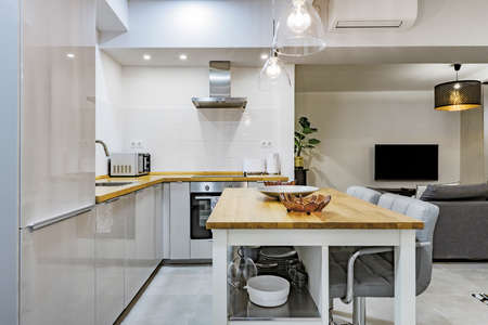Image of a kitchen with solid wood countertops, stainless steel appliances, clear lampshades, a matching island with tall gray chairs, and a living room with a TV and air conditioningのeditorial素材