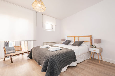 Bedroom with a large double bed with a wooden headboard, wooden nightstands and a white drawer, a gray blanket and matching cushions and windows with white blindsのeditorial素材