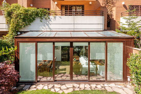 Covered terrace with a brown aluminum structure and glass with access to the house on the ground floor and a garden with a path of stone slabs and lawnのeditorial素材