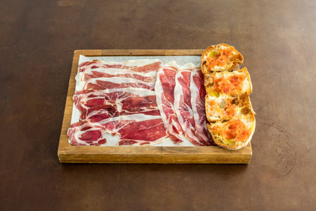 Wooden tray with a portion of typical Spanish acorn-fed ham with a slice of tumaca bread with olive...