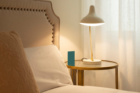 Corner of a bedroom with beige fabric upholstered headboard, gold metal and glass nightstand with...