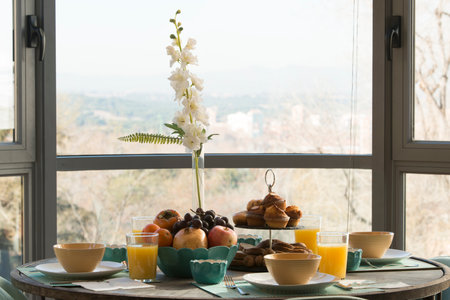 Round wooden dining table with blue placemats and breakfast service with a tray of assorted fruit, assorted scones, glasses of orange juice and a vase of artificial plantsの写真素材