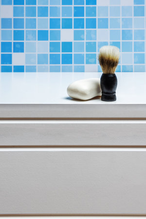 A barber's foam brush next to a bar of white soap in a bathroom with blue tiles and white cabinets