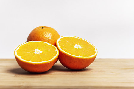 Two oranges bursting with vitamin c, one cut in half and one whole on a bamboo board with a white wall backgroundの写真素材