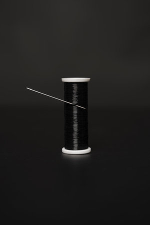 A lone spool of shiny black thread with a pricked sewing needle on a black surfaceの写真素材