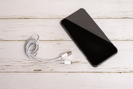 Phone with carbon fiber case and coiled white charging cable on white wooden tableの写真素材