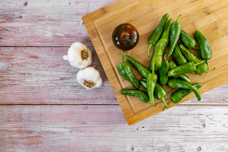 Fresh Galician green peppers, a ripe kumato tomato and a few heads of garlic on a bamboo board and a wooden tableの写真素材