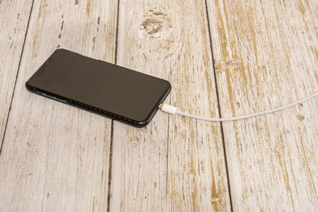 A black smartphone stuck with a white charging cable plugged into the phone on a wooden tableの写真素材