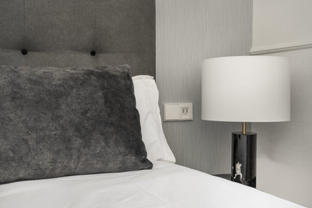 Detail of a bedroom with a gray fabric headboard, a gray velvet cushion and a lamp with a white...