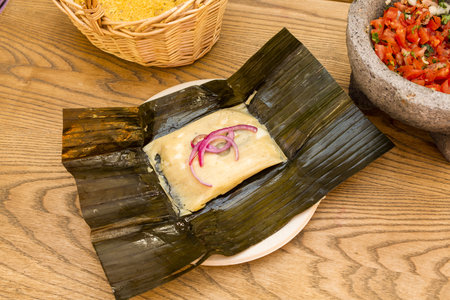 Mexican tamale of cochinita pibil stewed inside corn cob leaf with volcanic stone molcajete filled with pico de galloの写真素材